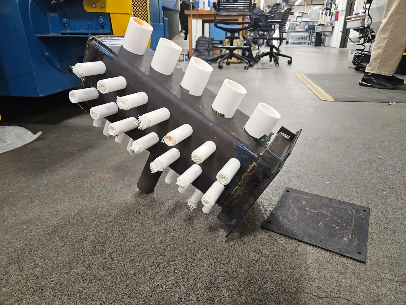Steel sample mold with rows of short white PTFE tubes packed with vent media, sitting on a shop floor next to a rotational molding machine